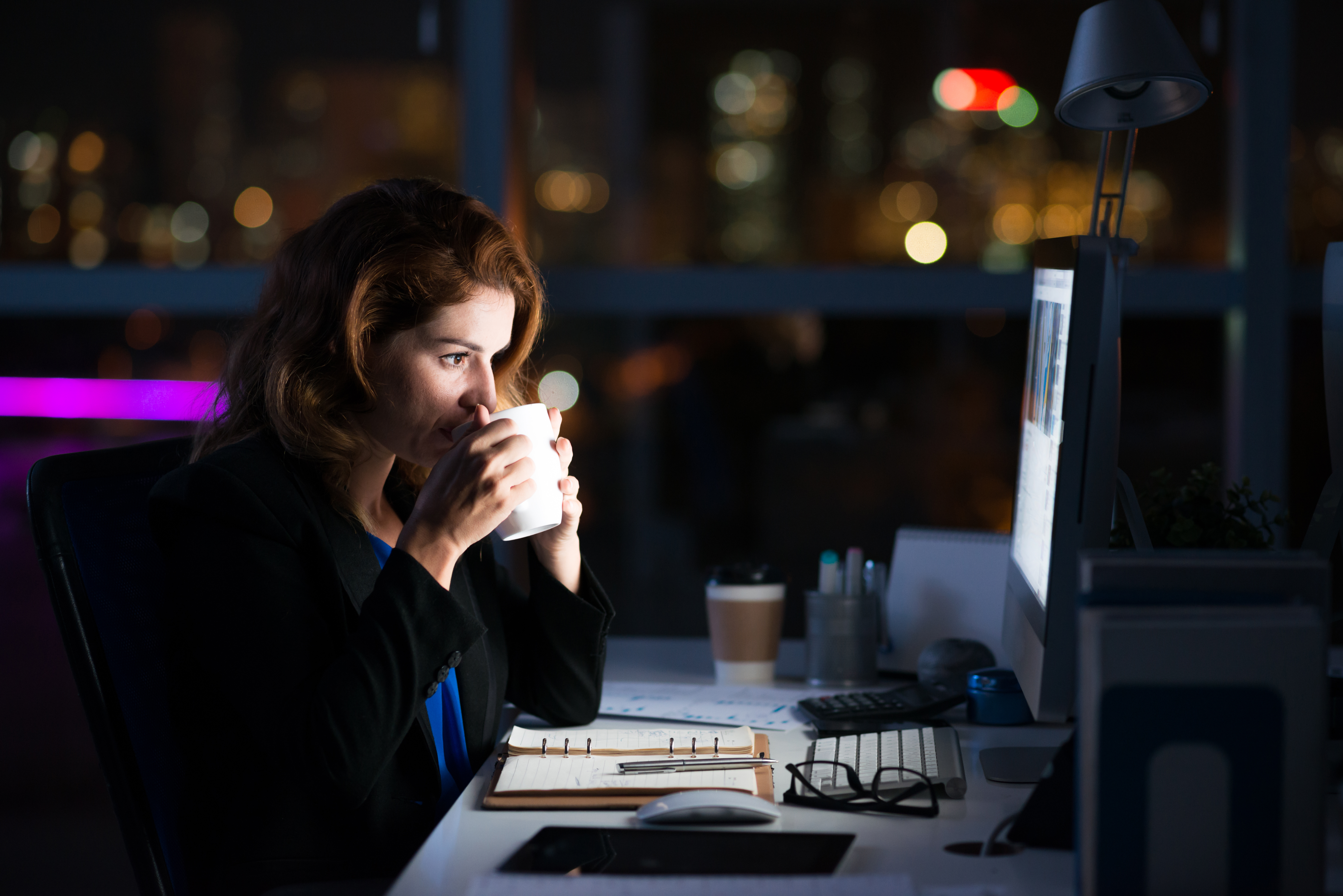 Business woman sitting at her work desk at night drinking coffee and staring at a computer screen symbolizing that she is working overtime.