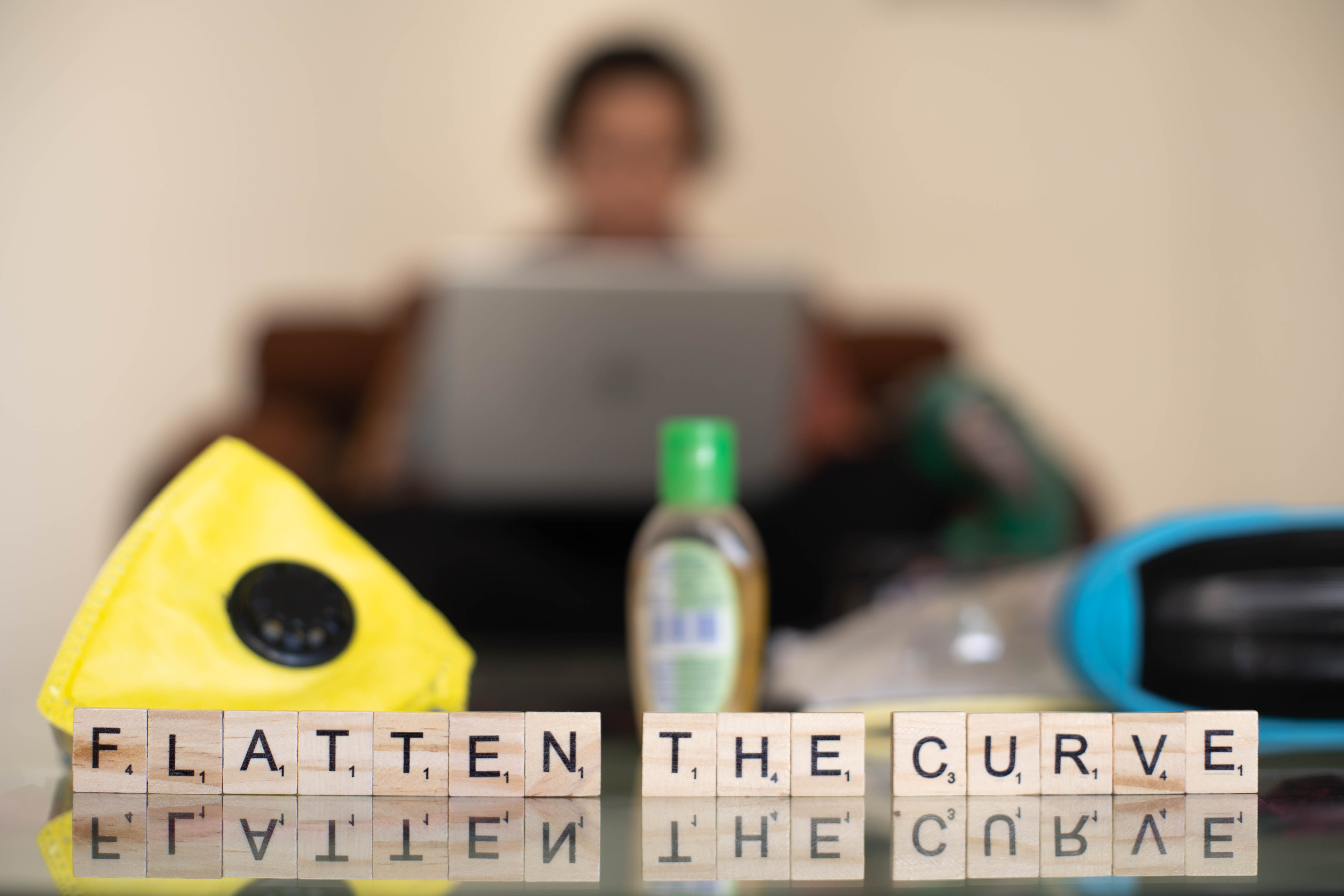Wooden blocks with flatten the curve with an out of focus background of a young indian girl woman working on her phone laptop while working from home on a self imposed quarantine in the corona covid 19 pandemic