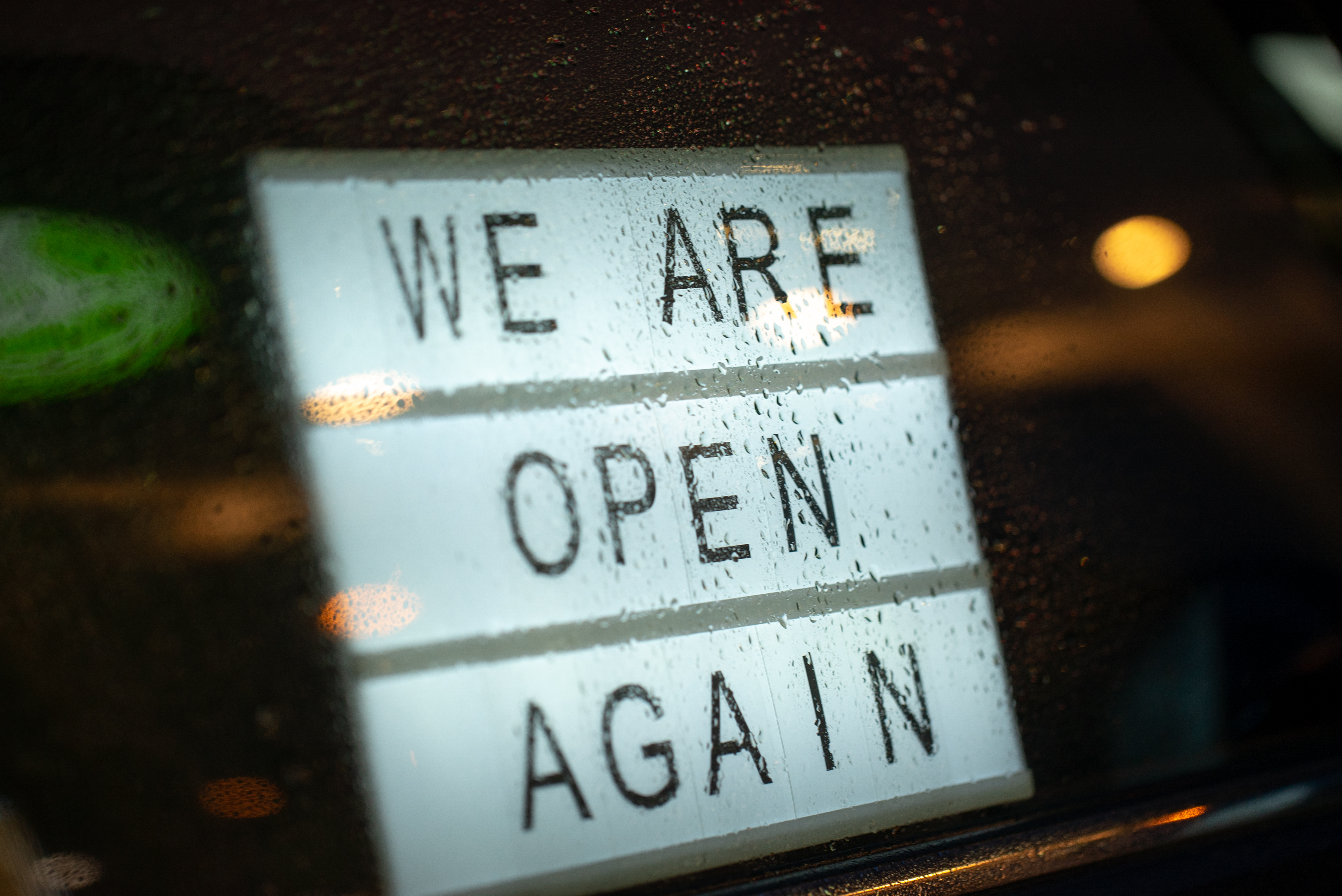 Lightbox with a sign we are open again behind a glass door of the cafe during the rain in the evening. We're open again after quarantine, photo of small business owner. Please wear a face mask and keep your distance to protect
