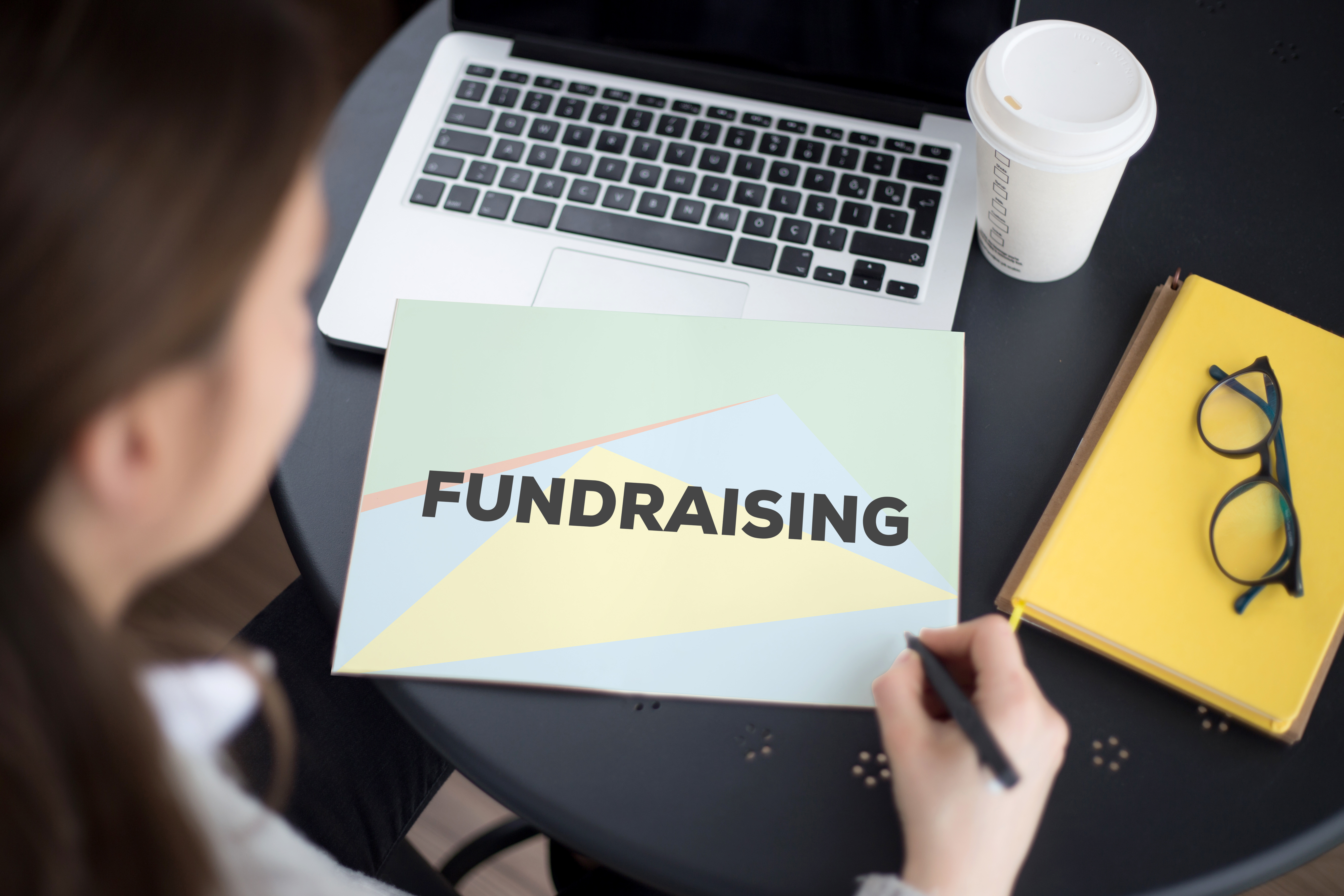Woman sitting at a desk looking down at a paper that says "fundraising" with laptop and coffee representing nonprofit legal issues. 