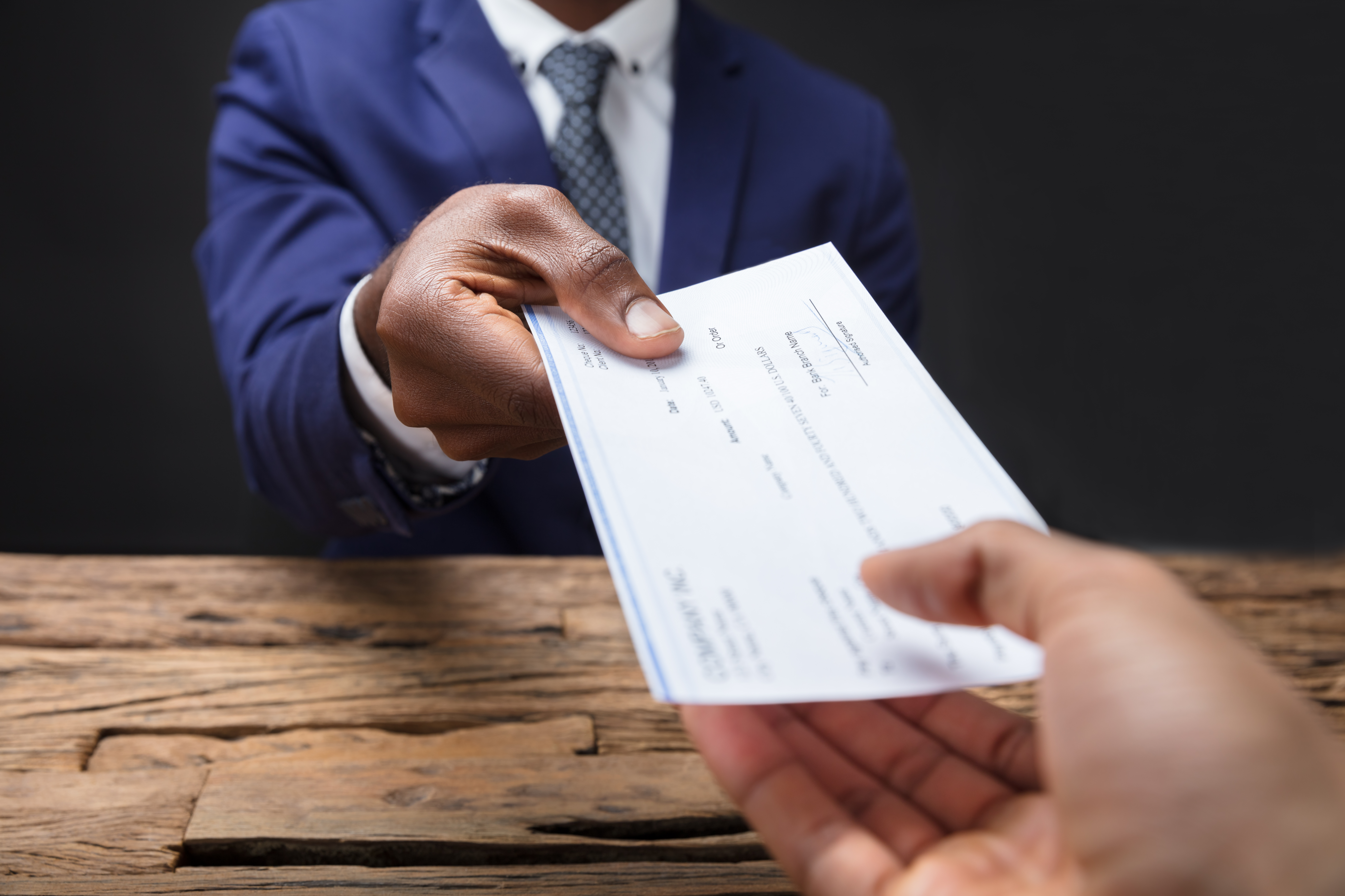 Close-up Of A Businessman's Hand Giving Check To Someone Over Wooden Desk Representing Counterfeit Check 