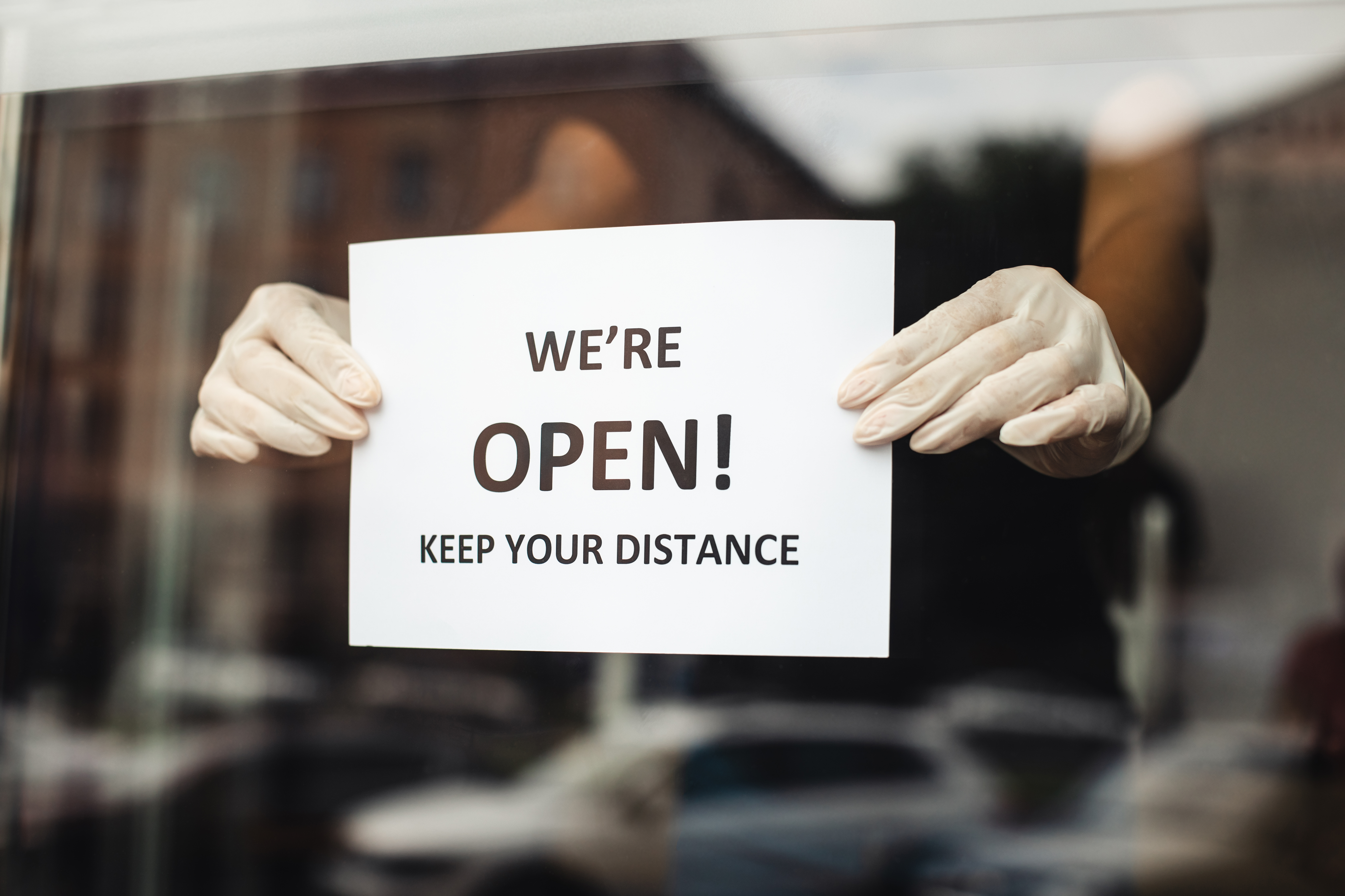 Woman in a storefront window hangs up a sign that says "we're open! Keep your distance" to represent businesses open with COVID restrictions in place. 