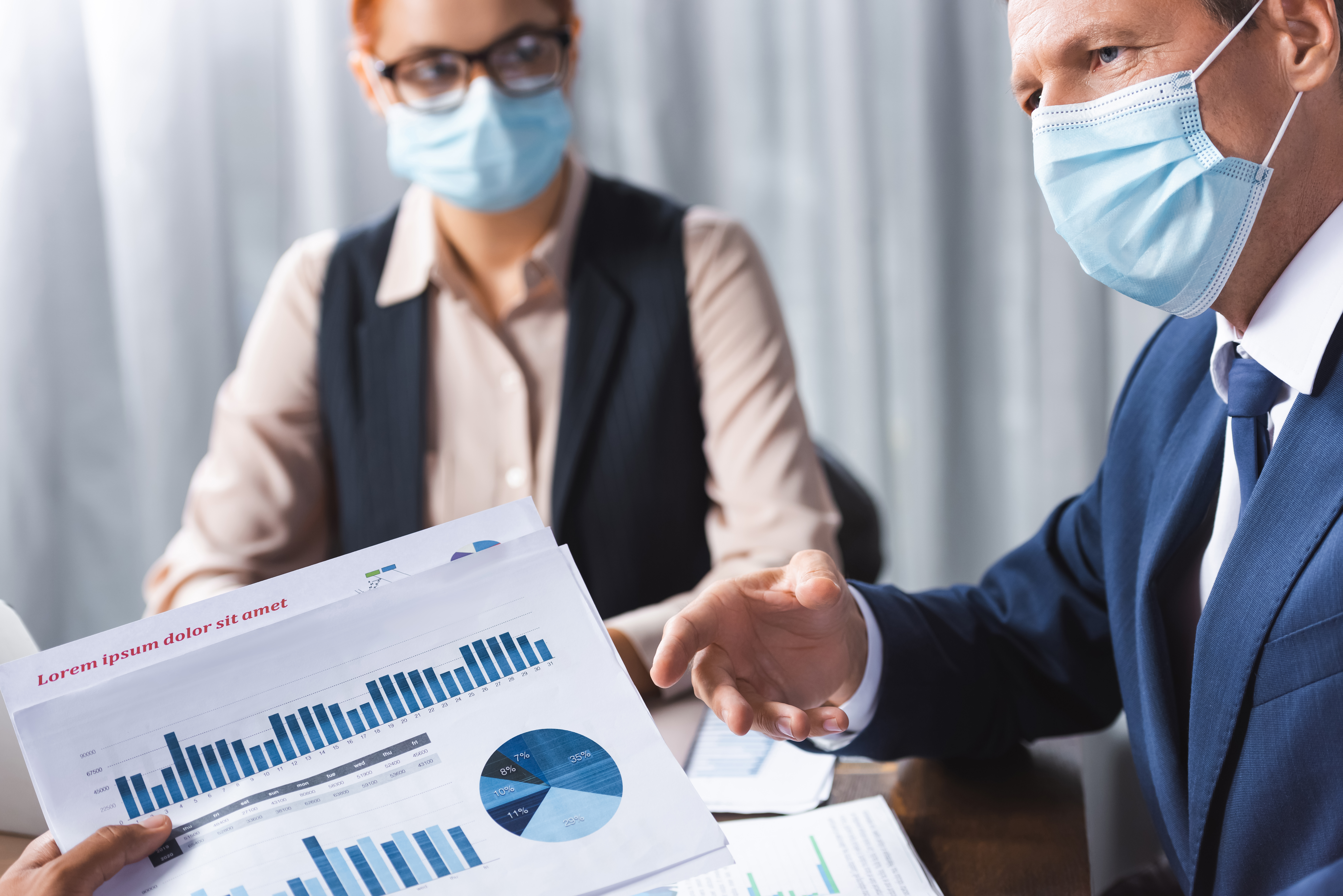 Businessman in mask next to colleague in mask gesturing as a 3rd collegue holds up graphs and charts of business data signifying employment law issues and COVID-19. 