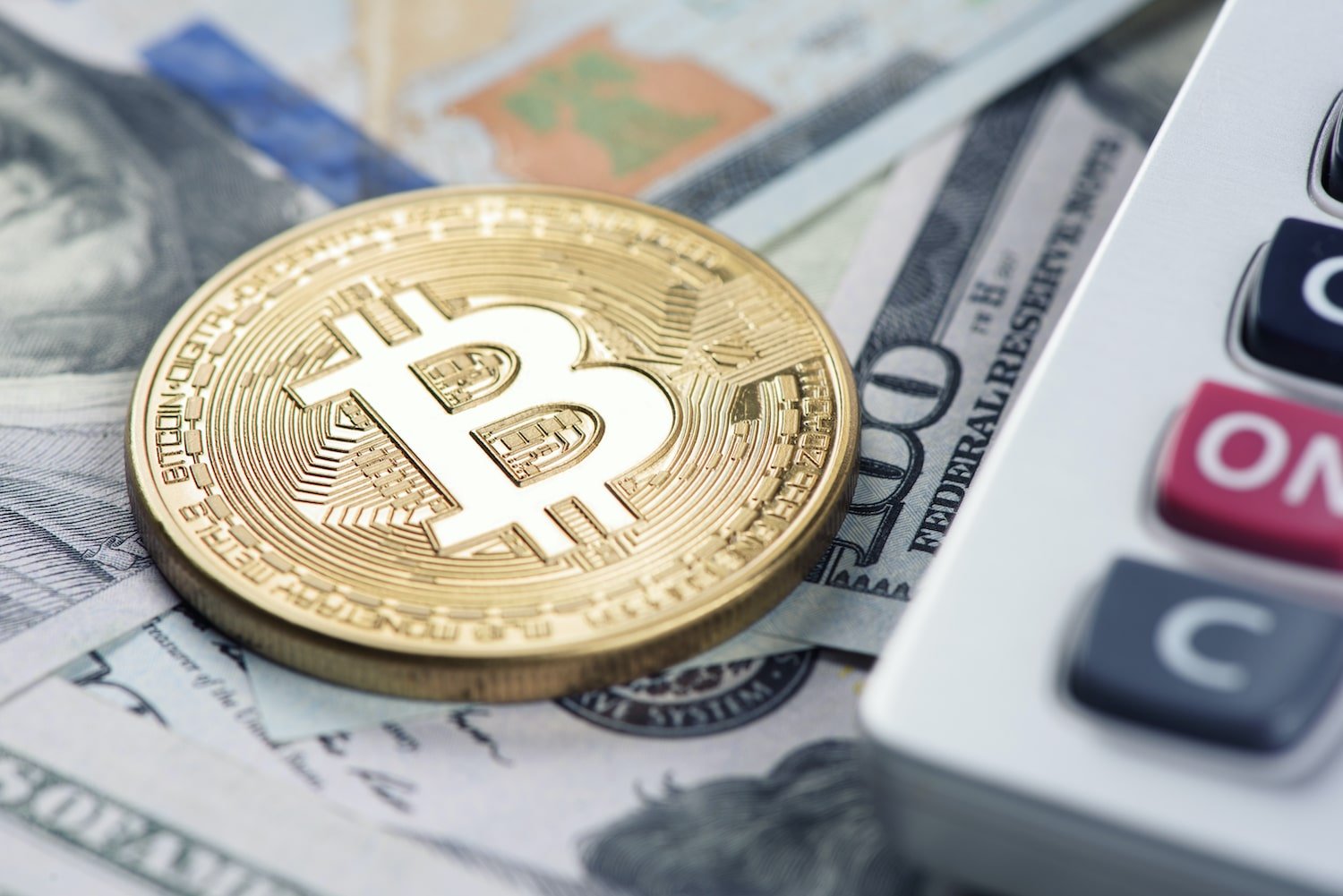 Image of a Bitcoin on top of a stack of cash next to a calculator.